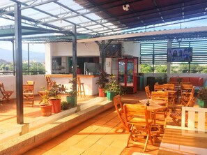 An outdoor patio with wooden tables and chairs, potted plants, and a red Coca-Cola refrigerator. - Bacano Hostel (Santa Marta)