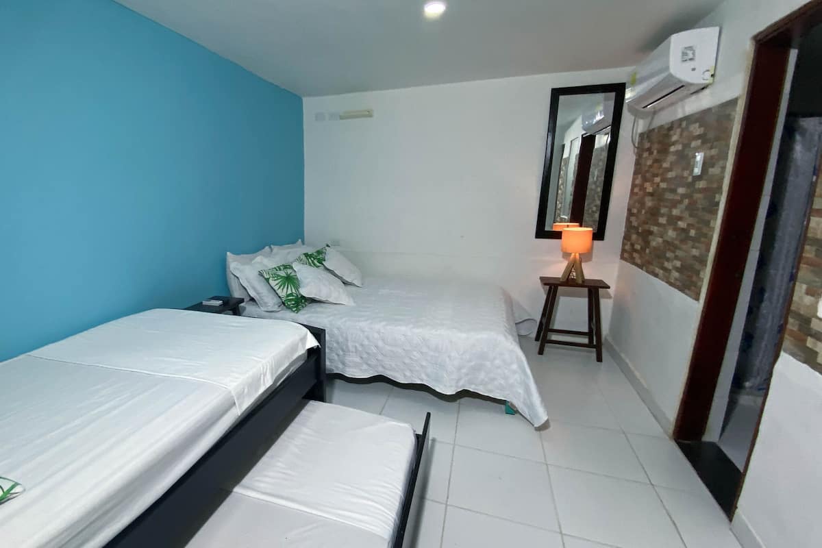 A small, clean room with two single beds, a bedside table with a lamp, and a wall-mounted air conditioner.