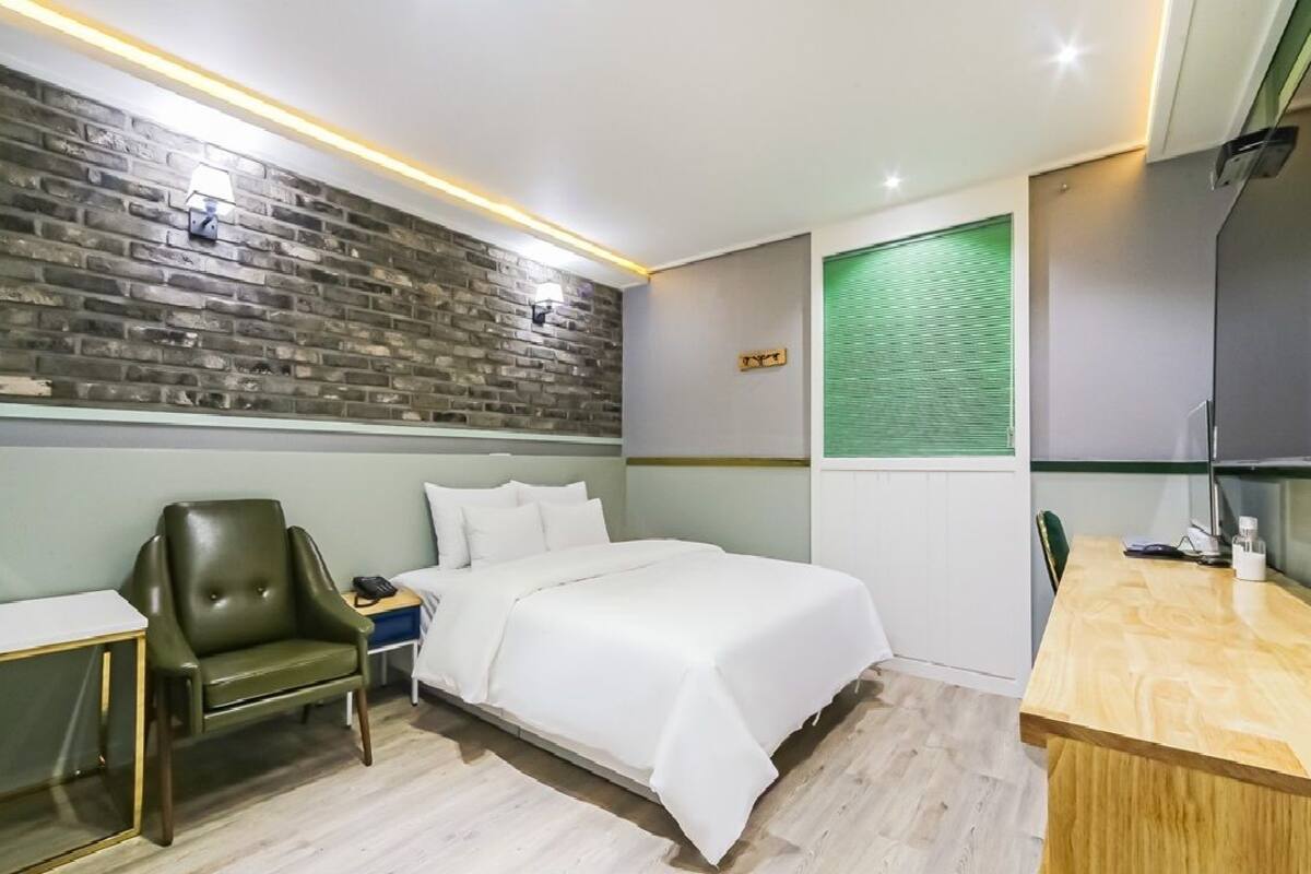 A modern hotel room with a brick-patterned accent wall, a bed with white bedding, a leather armchair, a wooden desk, and a green window blind.