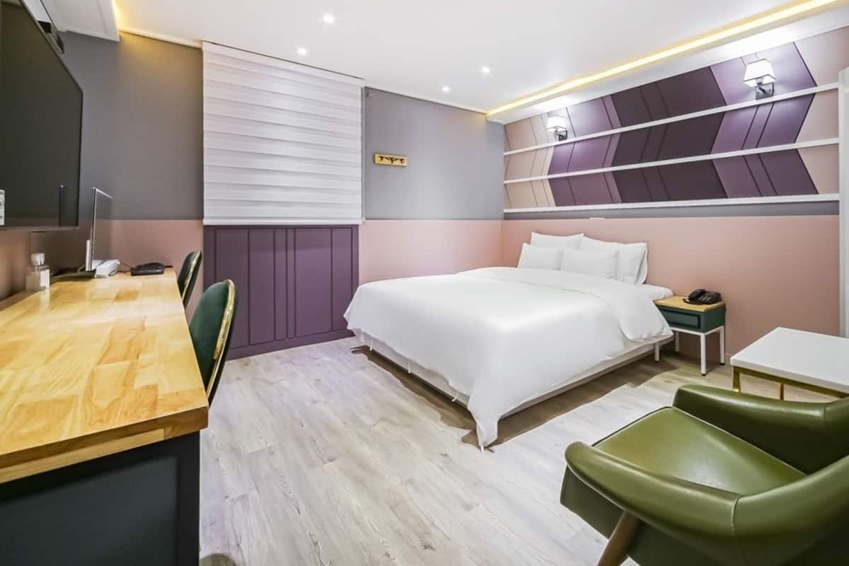 A modern hotel room with a wooden desk, a bed with white linens, a green armchair, and a bedside table with a lamp.