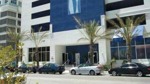 A multi-story hotel with a prominent blue and white facade, a parking lot with several cars, and palm trees in the foreground.