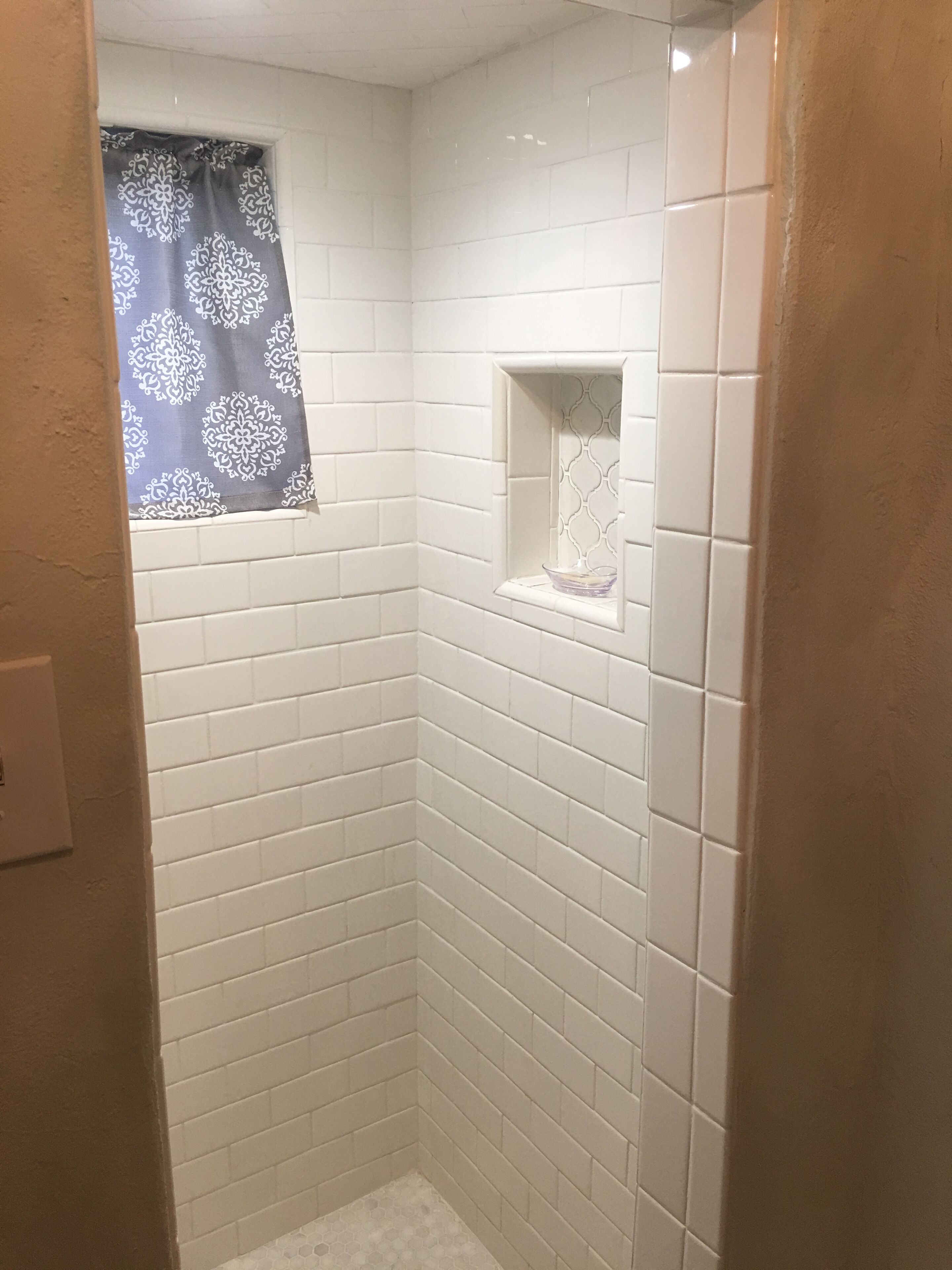 A small, enclosed shower area with white tiles and a window with a patterned curtain.