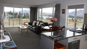 Una sala de estar moderna con un sofá de cuero negro, una mesa de comedor y una zona de cocina con un fregadero y estufa. Amplias ventanas ofrecen vistas a montañas y un lago.