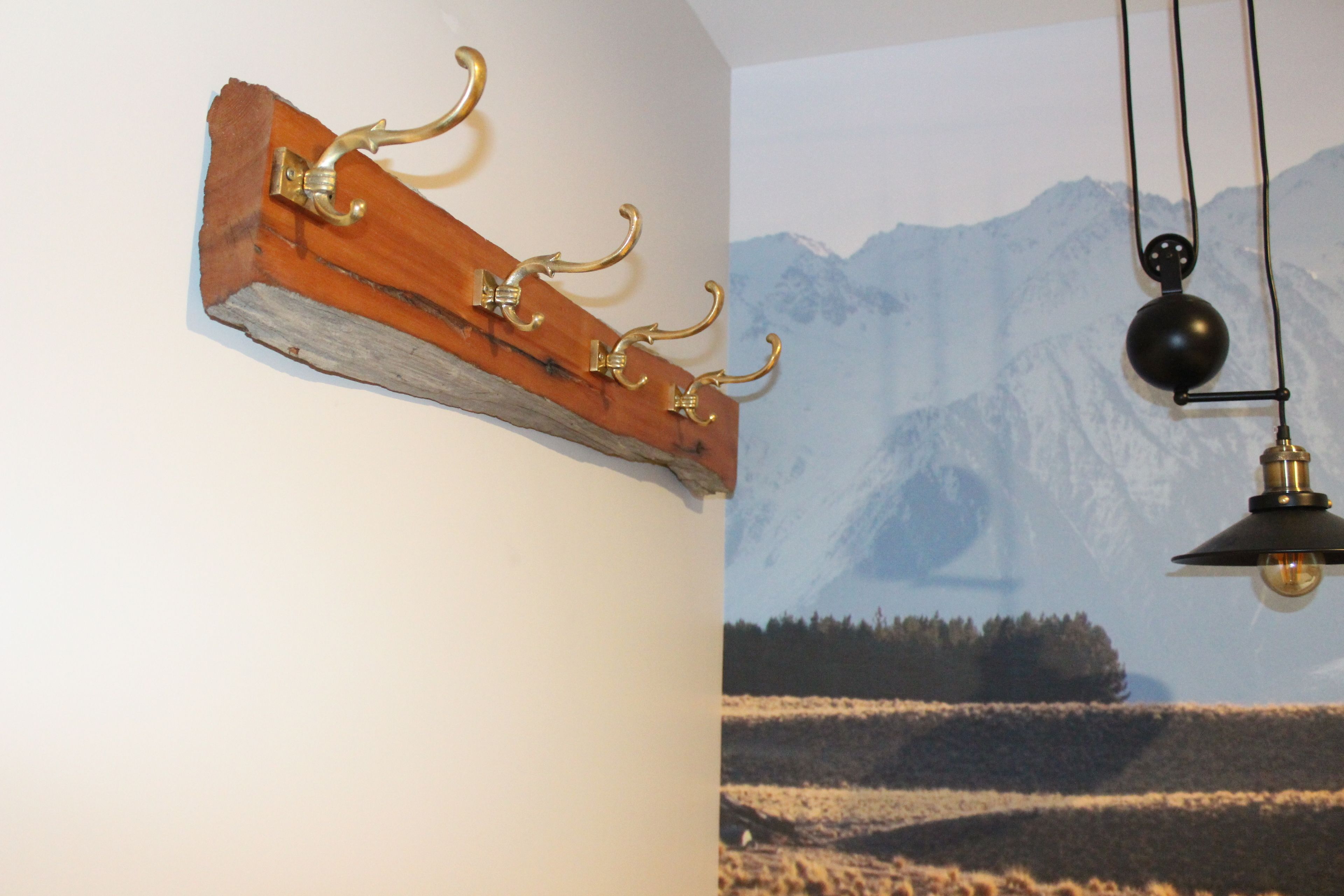 Een houten kapstok met messing haken voor een muur met een berglandschap op de muur.