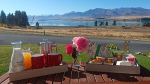 Un montaje para picnic con una botella de leche, un vaso de jugo de naranja, tazas rojas, un florero con flores rosadas y recipientes de comida sobre una mesa de madera.