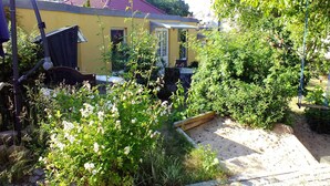Une maison jaune avec un toit rouge, entourée d’une végétation luxuriante et d  ’un jardin où poussent différentes plantes et fleurs.