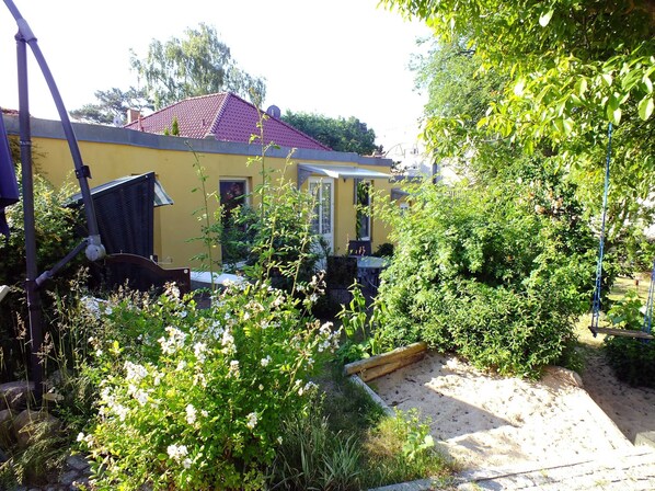 A yellow house with a red roof, surrounded by dense greenery and a garden area with various plants and flowers.