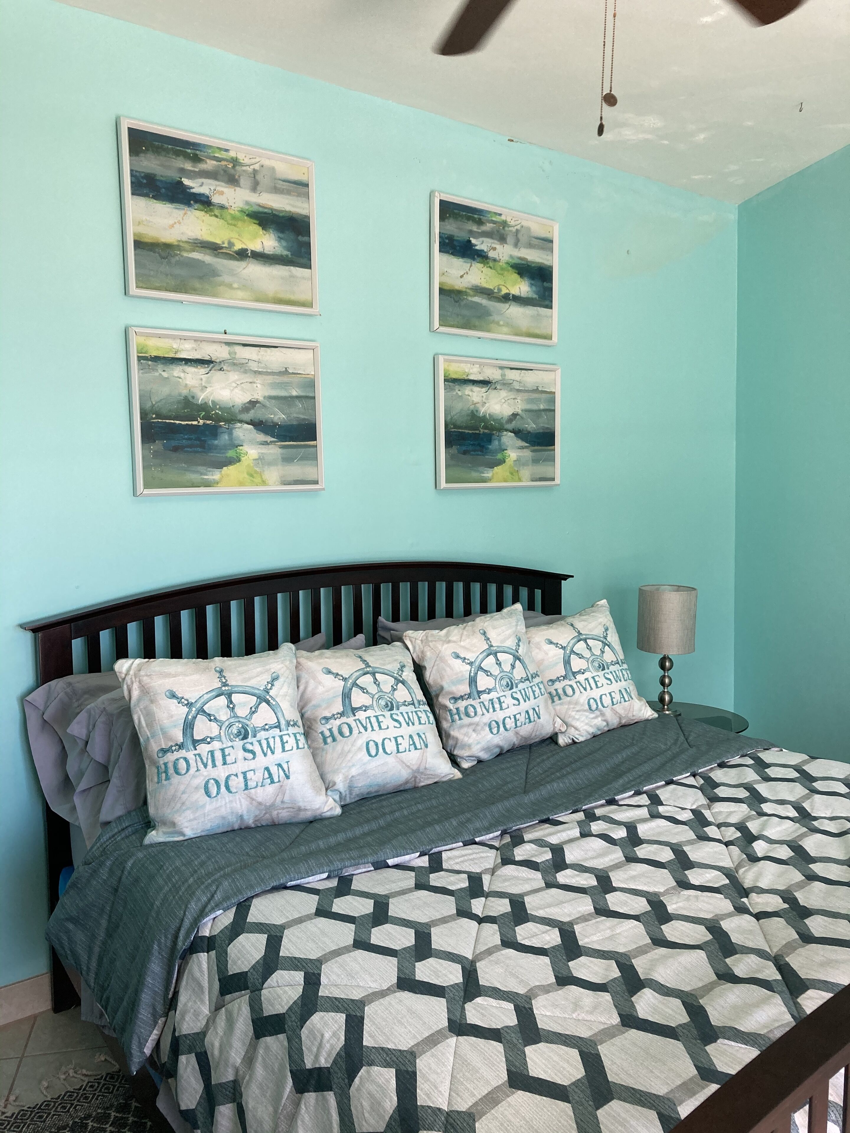 A bedroom with a bed, a headboard, a nightstand, a lamp, and four framed abstract paintings on the wall.