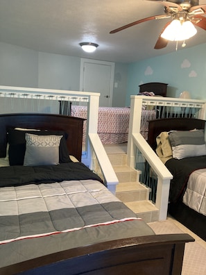 Two beds with wooden headboards and white railings, adorned with pillows and blankets, are set against a light blue wall with a cloud pattern.