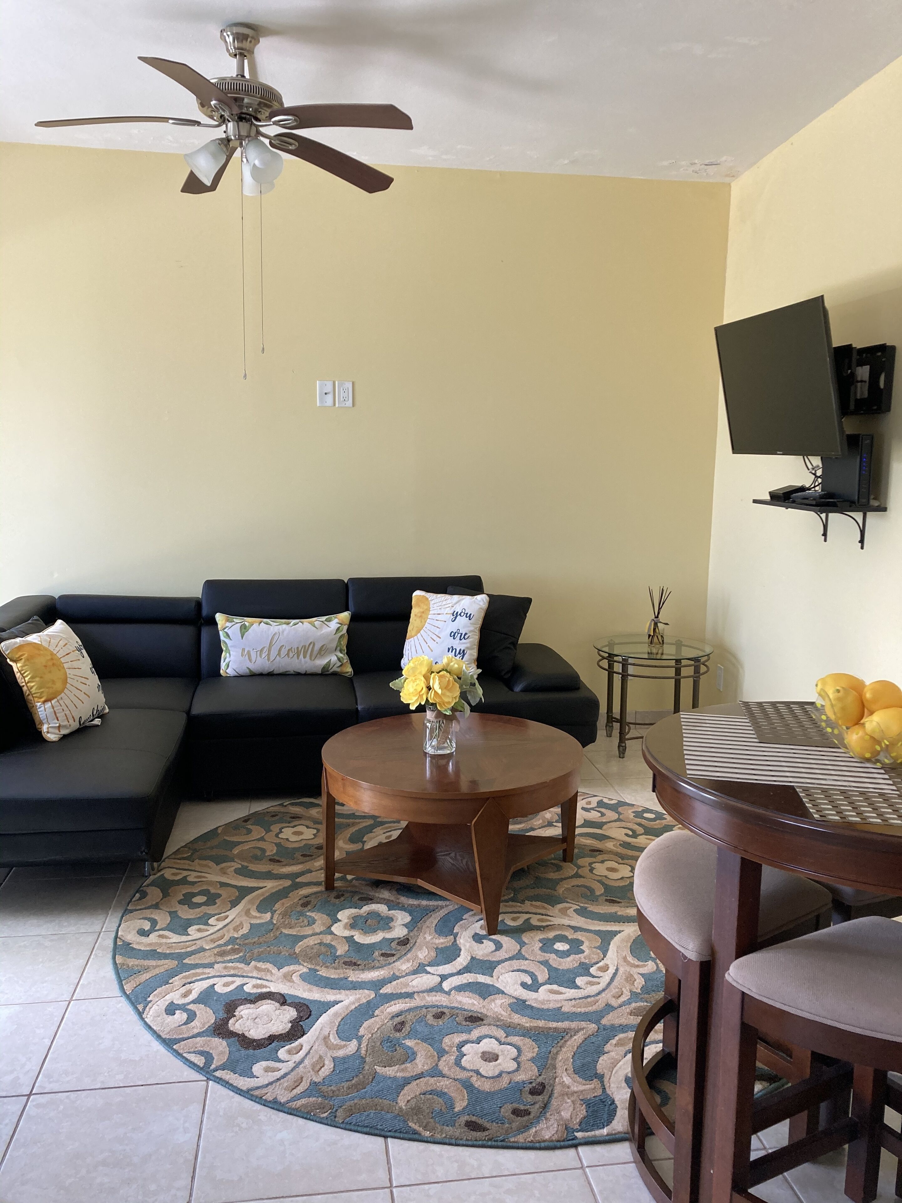 A living room with a black sofa, a coffee table with a vase of flowers, a patterned rug, a ceiling fan, and a mounted TV.