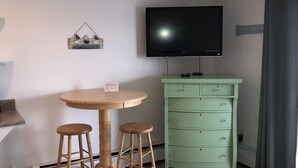 Een kleine kamer met een ronde houten tafel, twee houten krukken, een groene kast en een flatscreen tv aan de muur.