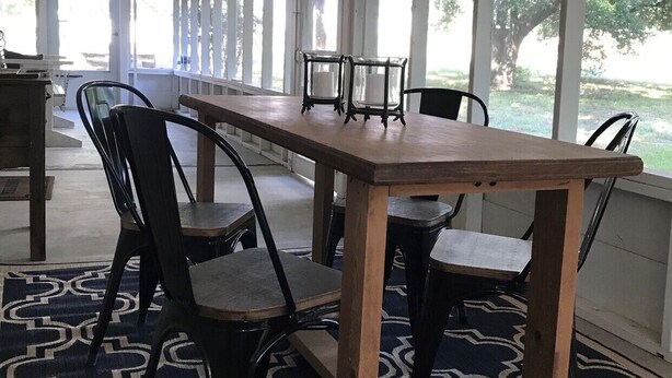 Un espacio exterior cubierto para comer con una larga mesa de madera y sillas, luces colgantes y grandes ventanales con vistas a un jardín.