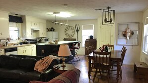 Un salón amplio con un sofá de cuero negro, un sofá de cuadros y una mesa de comedor de madera con sillas. Se ve en el fondo una zona de cocina con armarios blancos y un reloj grande.