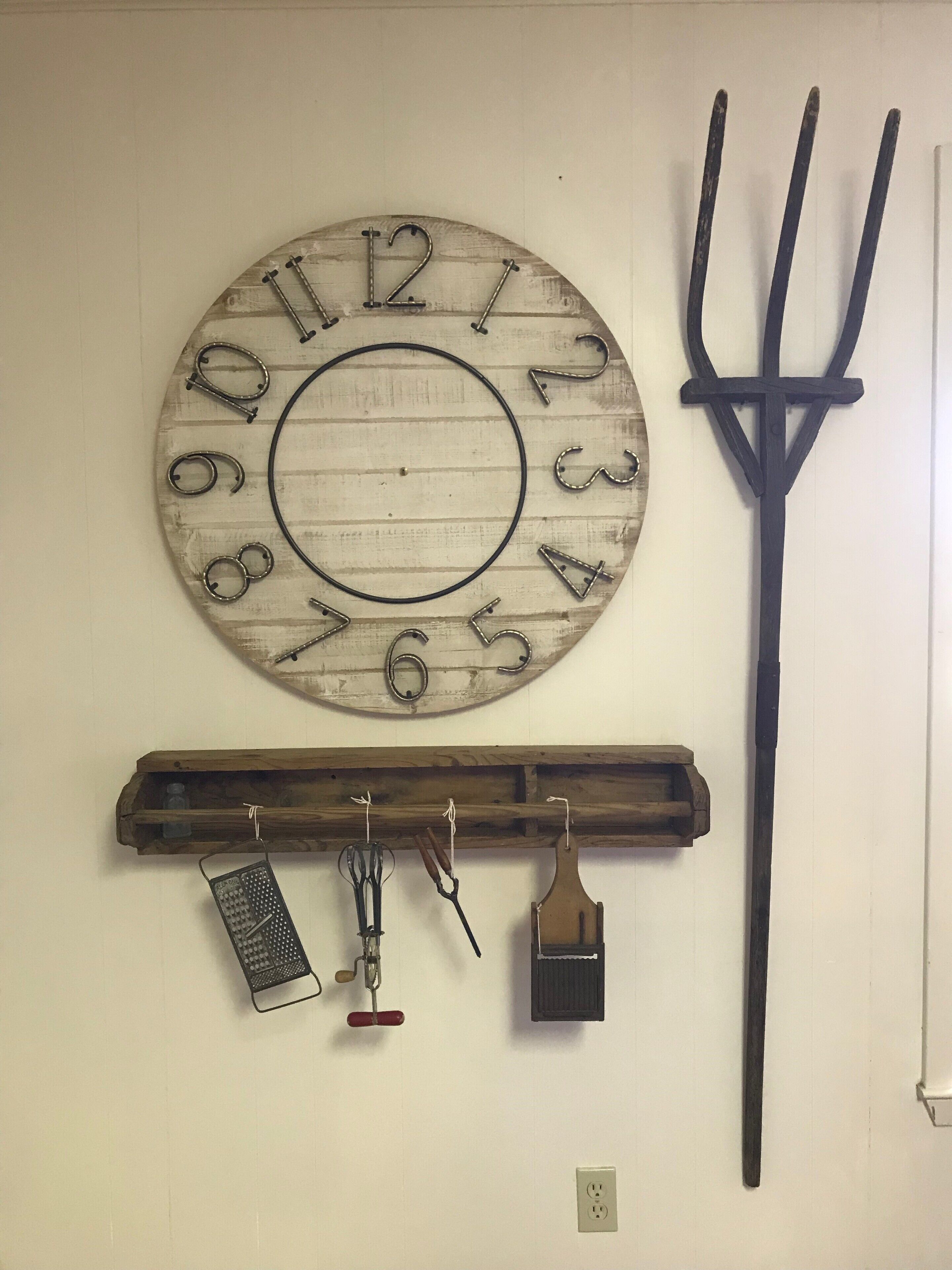 A rustic wall clock with numbers and hands, a wooden shelf with kitchen utensils, and a black pitchfork.