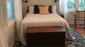Un dormitorio con una cama grande, lámparas de noche, un banco de madera, un tapete estampado y una ventana con cortinas.