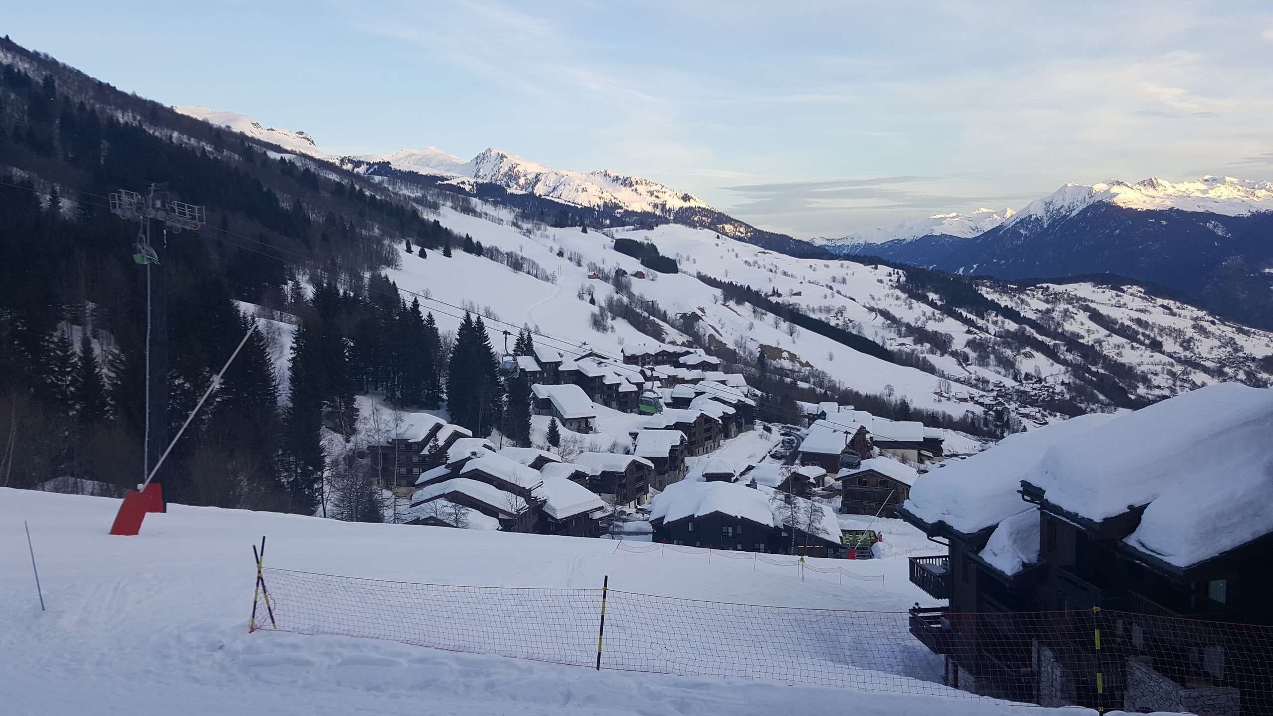 Un village de montagne enneigé, avec ses maisons traditionnelles en bois, ses remontées mécaniques et une vue dégagée sur les sommets environnants.