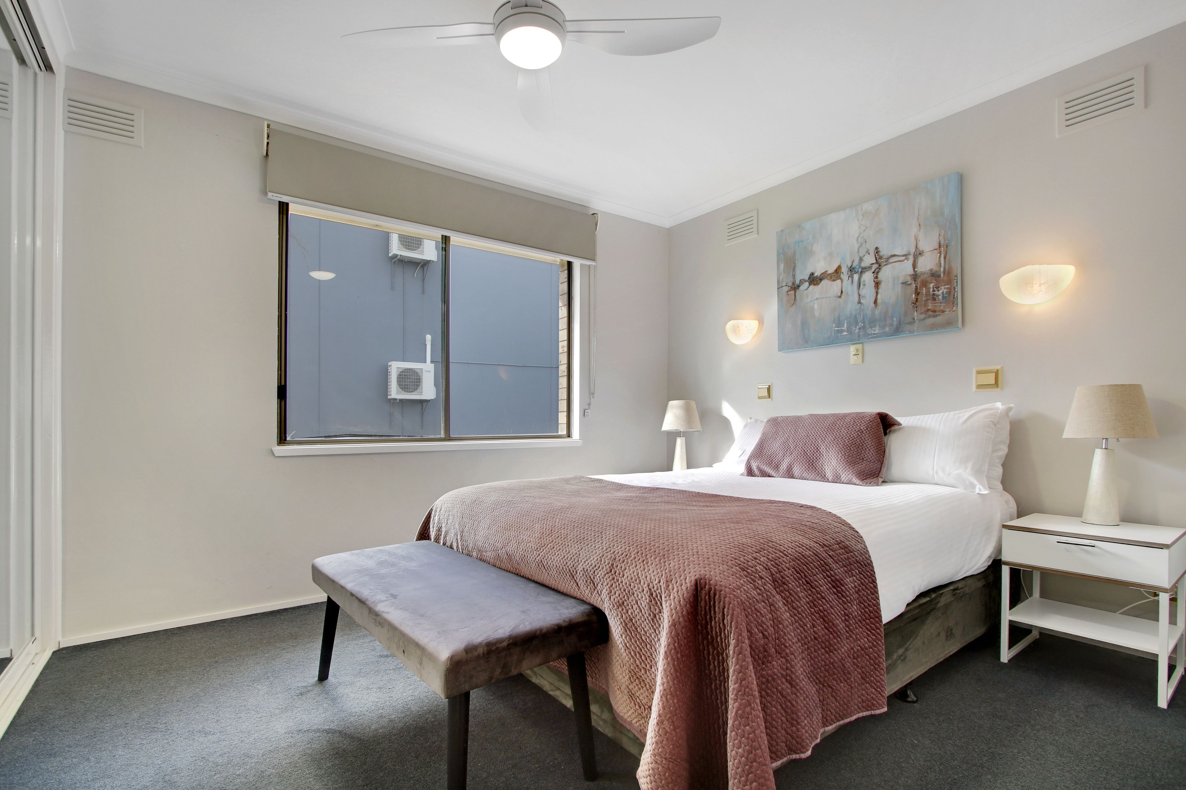 A bedroom with a bed, bench, bedside tables, a ceiling fan, and a window with air conditioning units.
