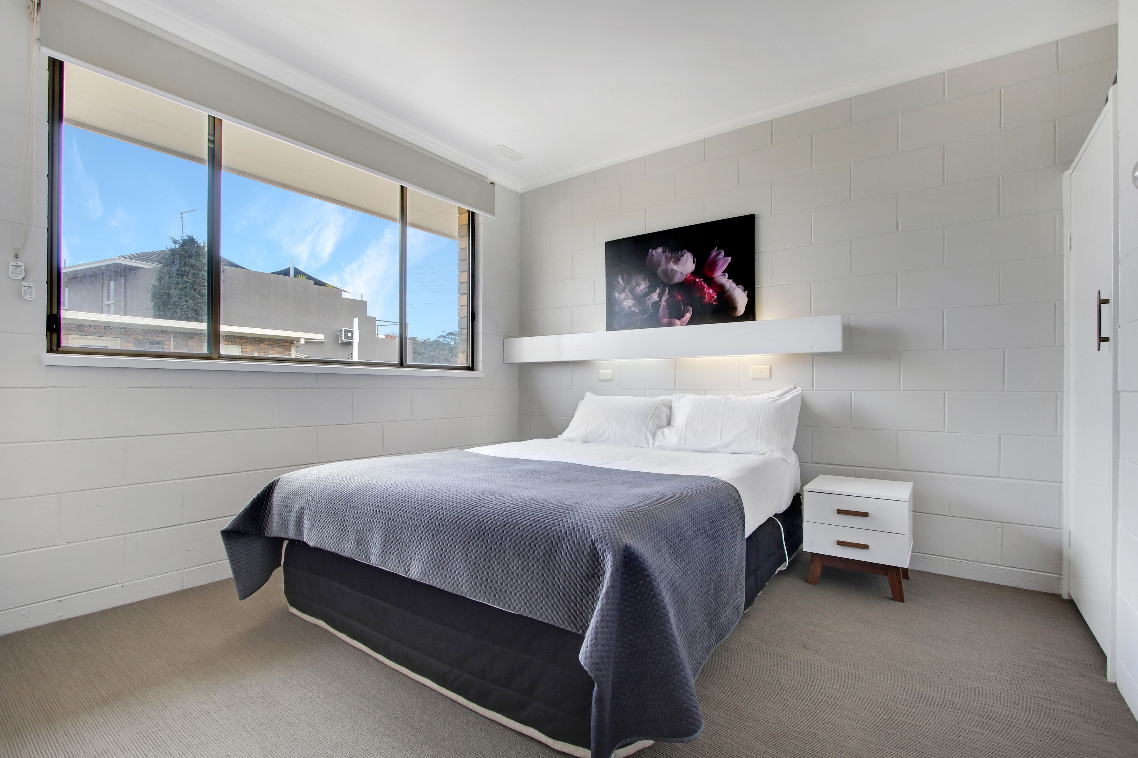 A bedroom with a bed, a nightstand, a window, and a wall-mounted artwork.