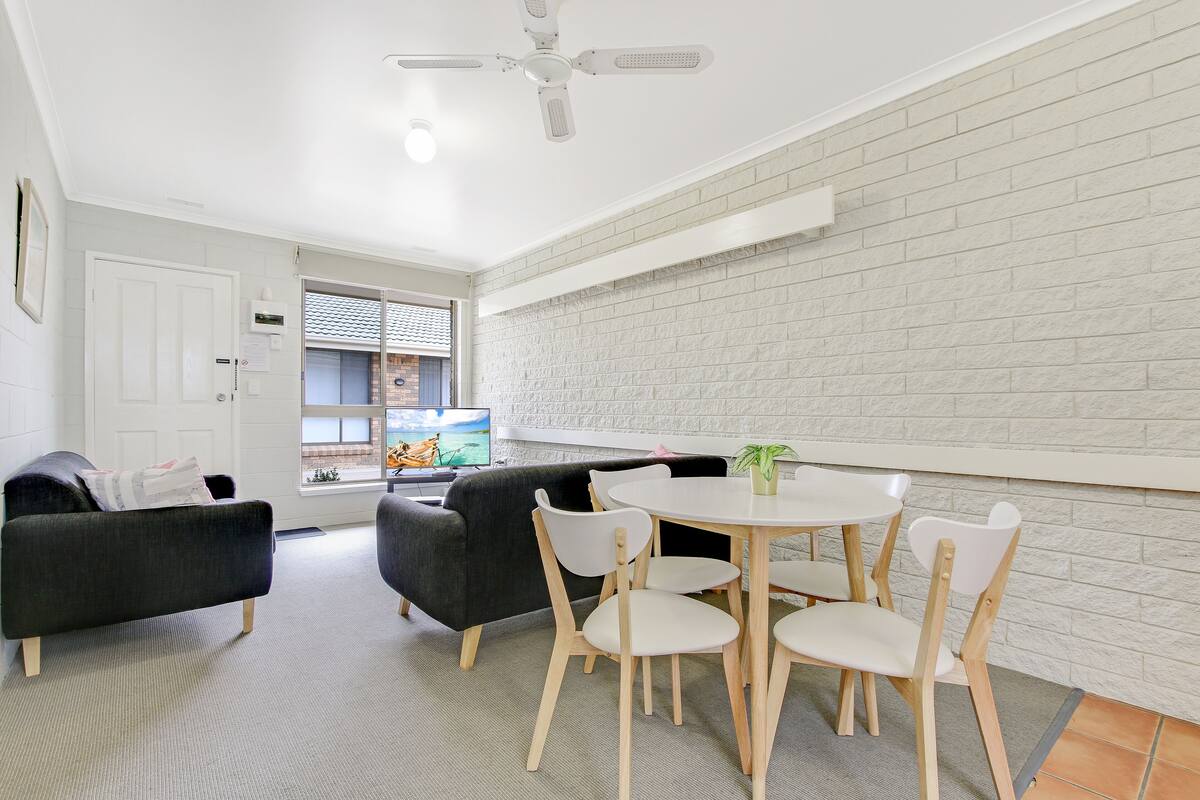 A modern living room with a white brick wall, a ceiling fan, a round dining table with four chairs, and a dark-colored sofa.