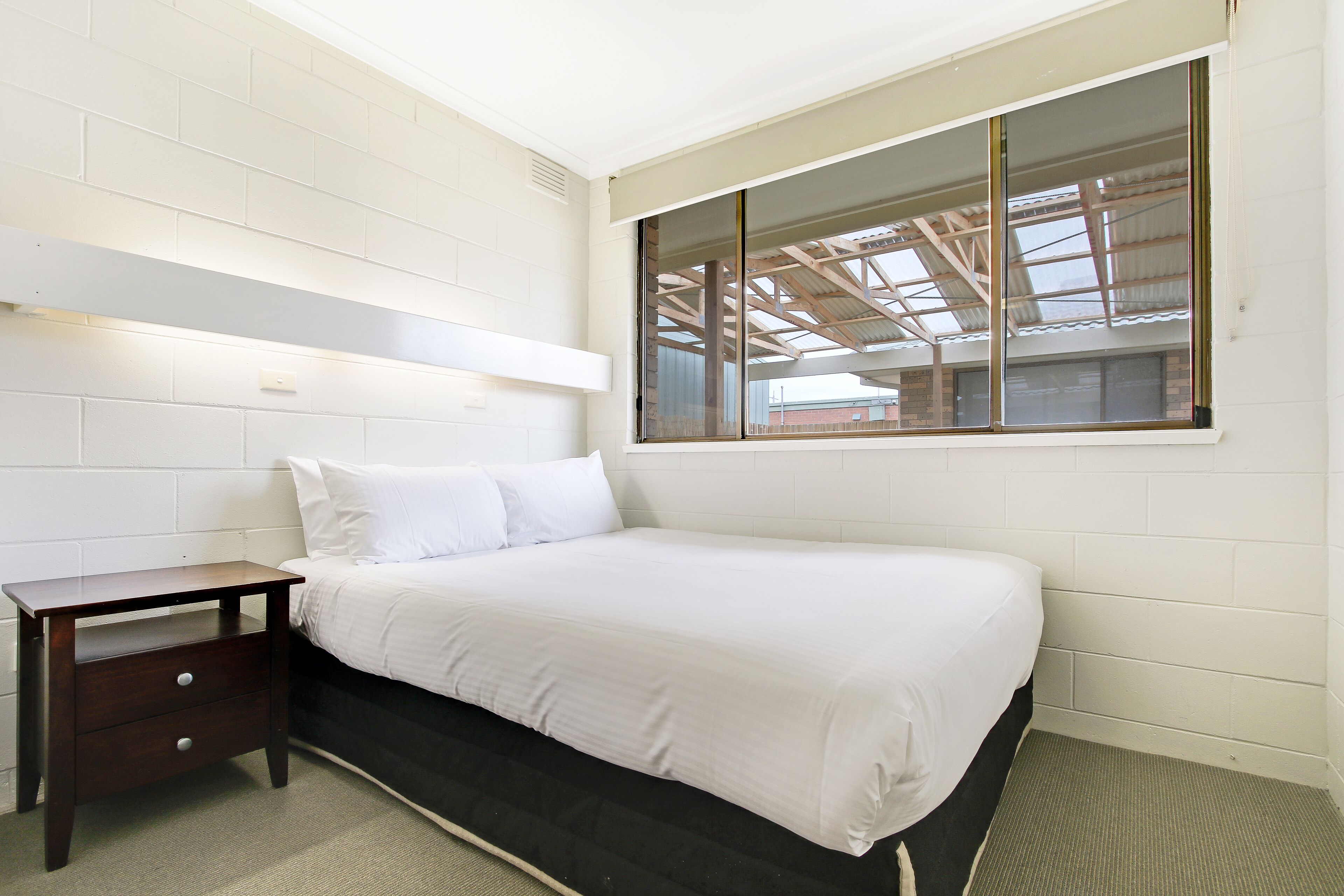 A bedroom with a bed, a nightstand, a window with a view of the roof, and white brick walls.