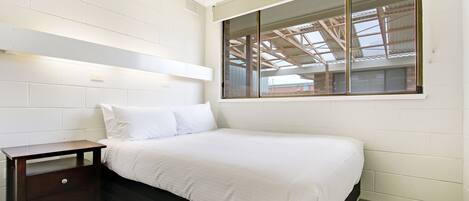 Un dormitorio con cama, mesita de noche, ventana y vista al tejado, y paredes de ladrillo blanco.