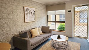 Una habitación con pared de ladrillo, un sofá con cojines, una mesa de centro redonda y una ventana con vistas a la vegetación.
