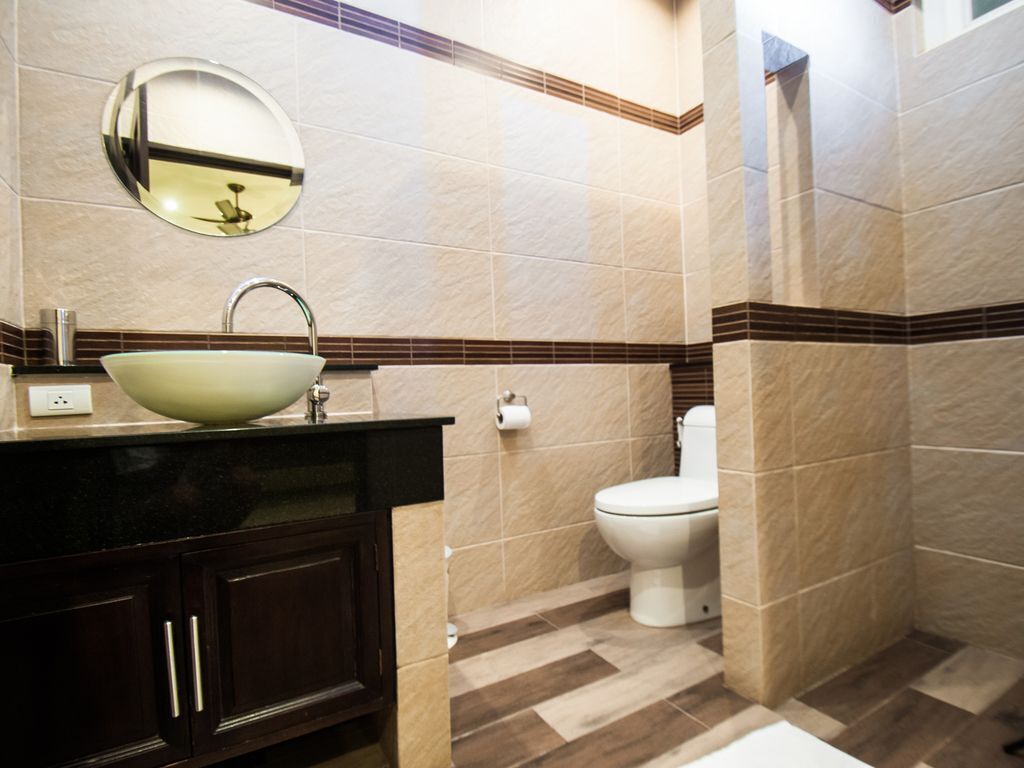Et badeværelse med en hvid vask, mørke træskabe, et rundt spejl og et toilet.
