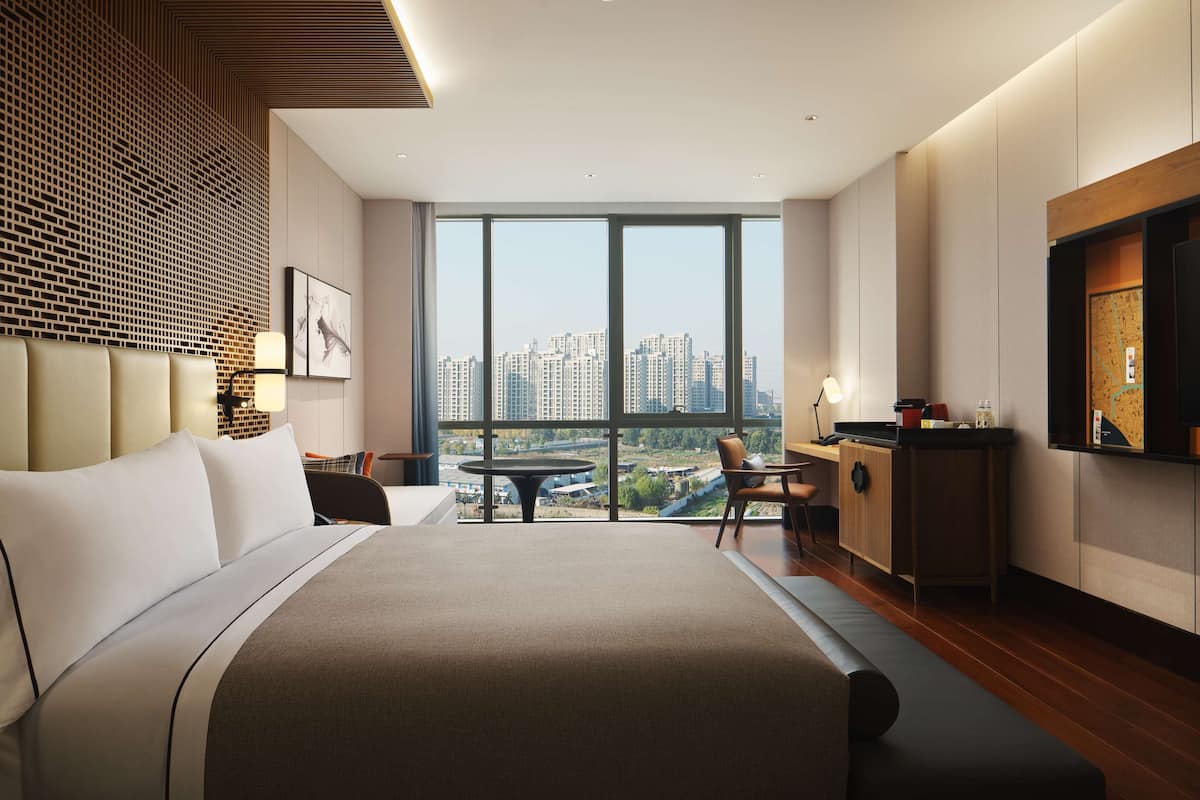 A modern hotel room with a large bed, a dining table, and a view of the cityscape.