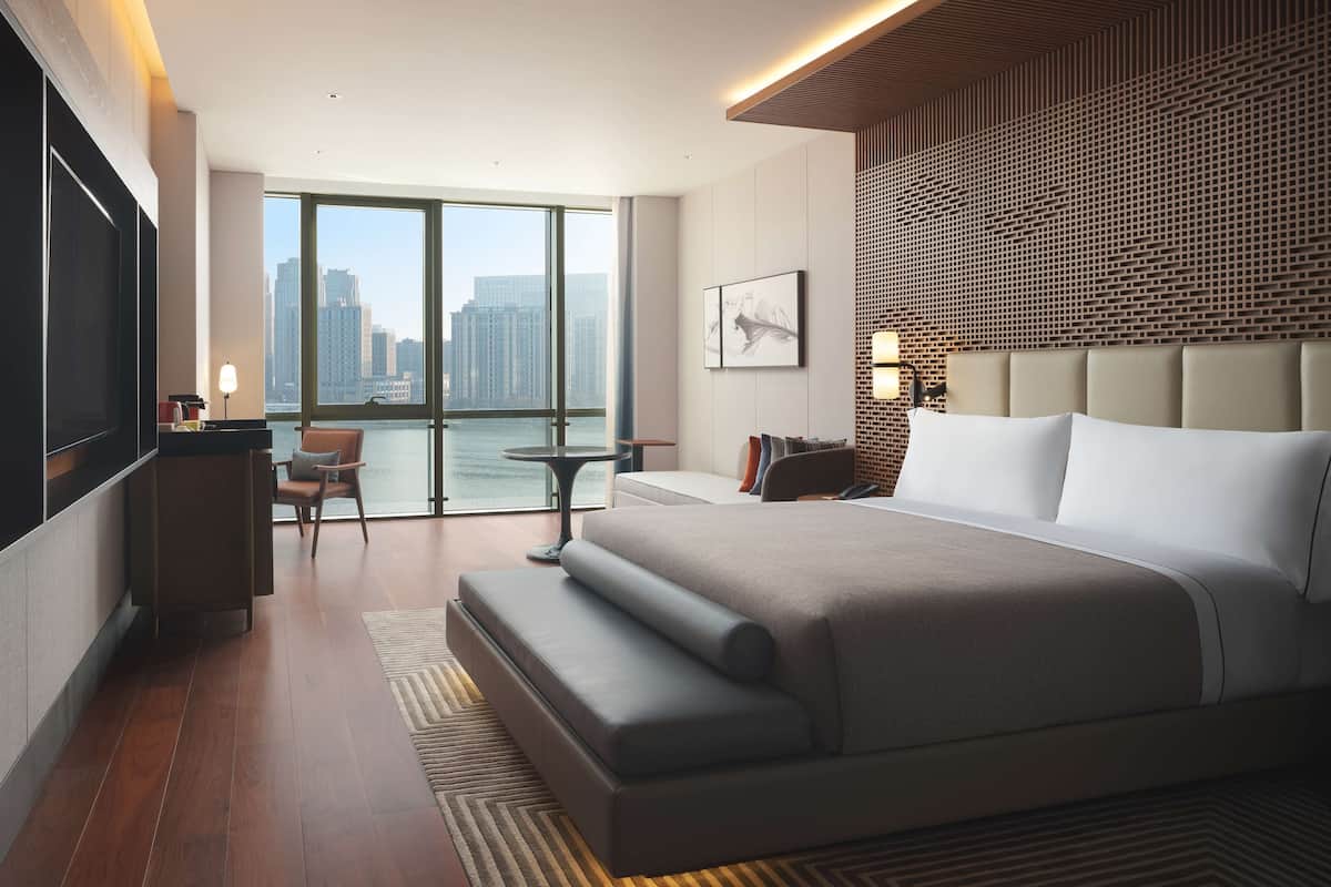 A modern hotel room with a large bed, a desk, a chair, and a view of the city.
