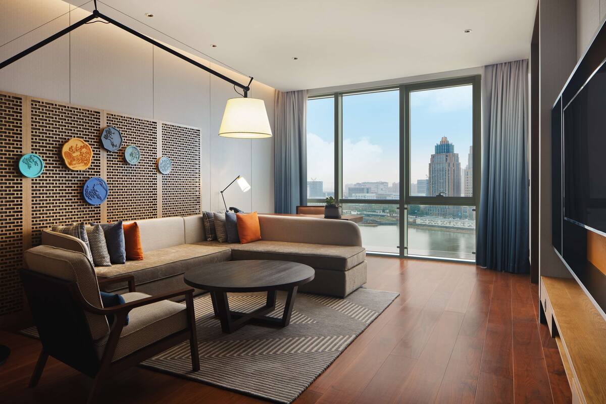 A modern living room with a sectional sofa, a coffee table, and a large TV. There is a city view through the window.