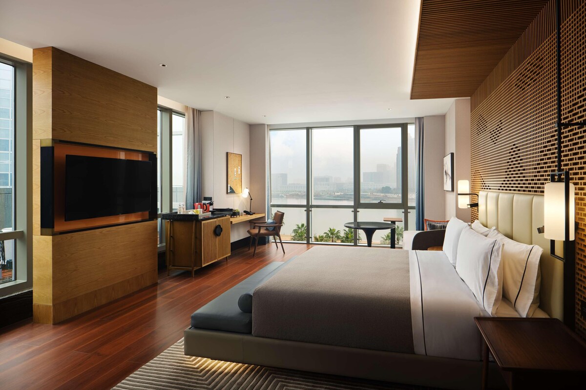 A modern hotel room with a large bed, a desk with a chair, a television, and a city view.