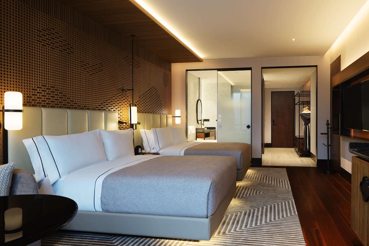 A modern hotel room with two beds, a TV, and a bathroom visible through a glass door.
