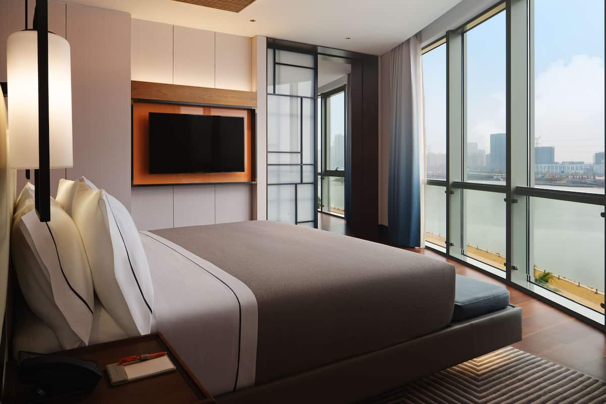 A modern hotel room with a large bed, a flat-screen TV mounted on the wall, and a view of a cityscape through large windows.