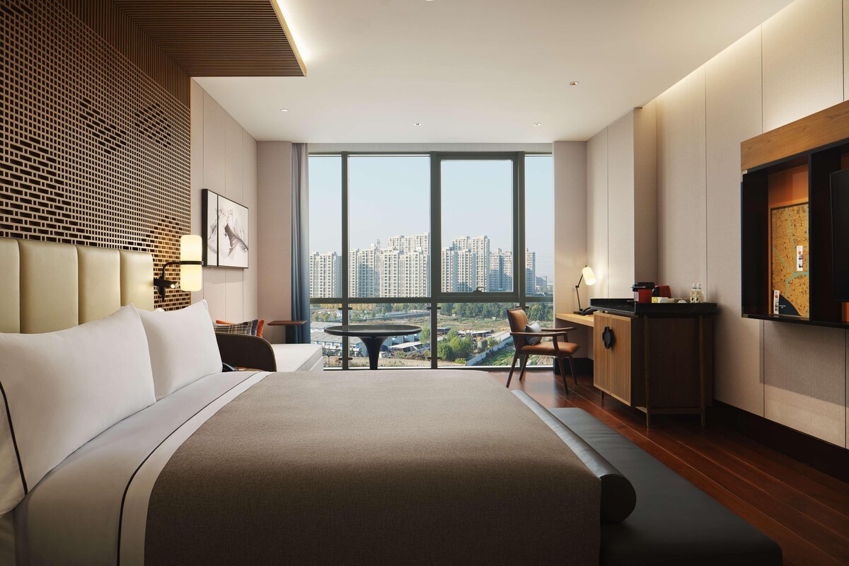 Una habitación de hotel moderna con una cama grande, una mesa de comedor, una zona de cocina y vistas a la ciudad a través de amplias ventanas.