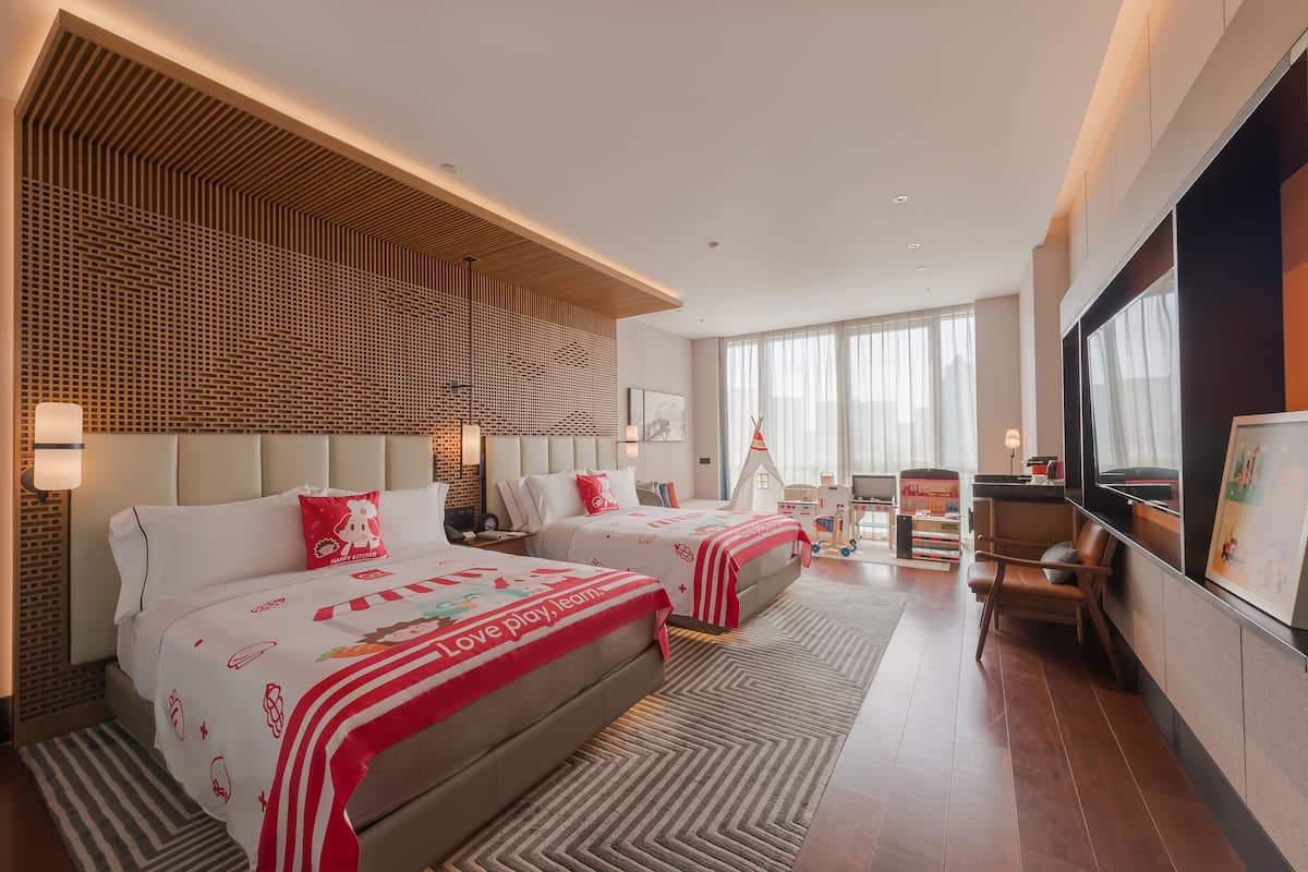 A hotel room with two beds, a large window, a TV, and a seating area with a chair and table.