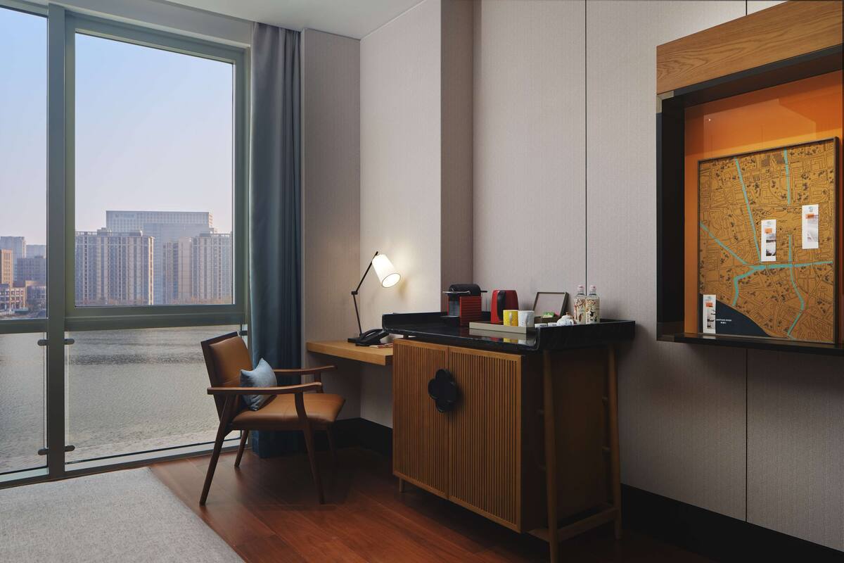 A hotel room with a desk, chair, and a large window offering a city view.
