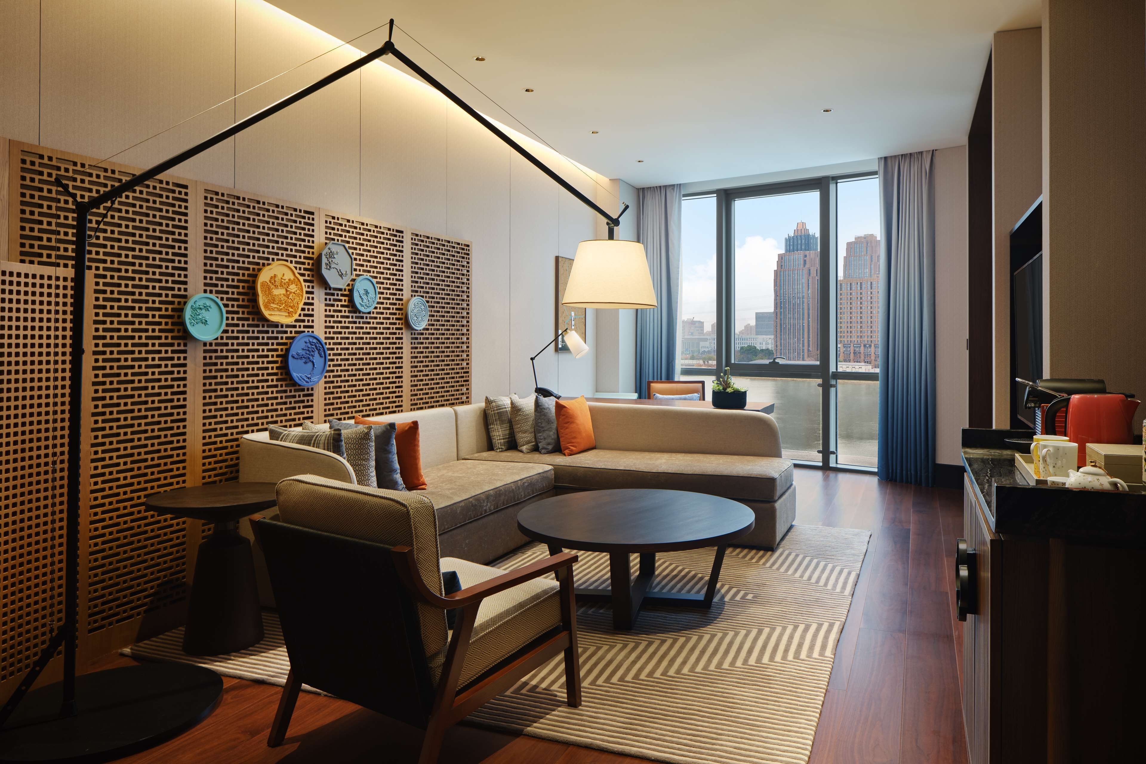 Una habitación de hotel moderna con sofá modular, una mesa de centro redonda, una lámpara de pie y vistas a la ciudad a través de ventanas amplias.