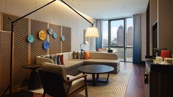 Una habitación de hotel moderna con sofá modular, una mesa de centro redonda, una lámpara de pie y vistas a la ciudad a través de ventanas amplias.