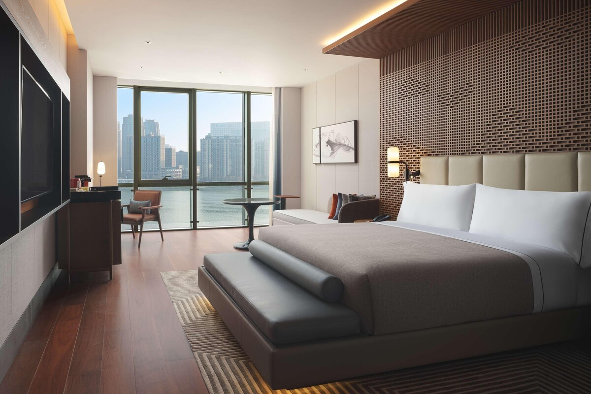 A modern hotel room with a large bed, a desk, a chair, and a view of the city skyline.