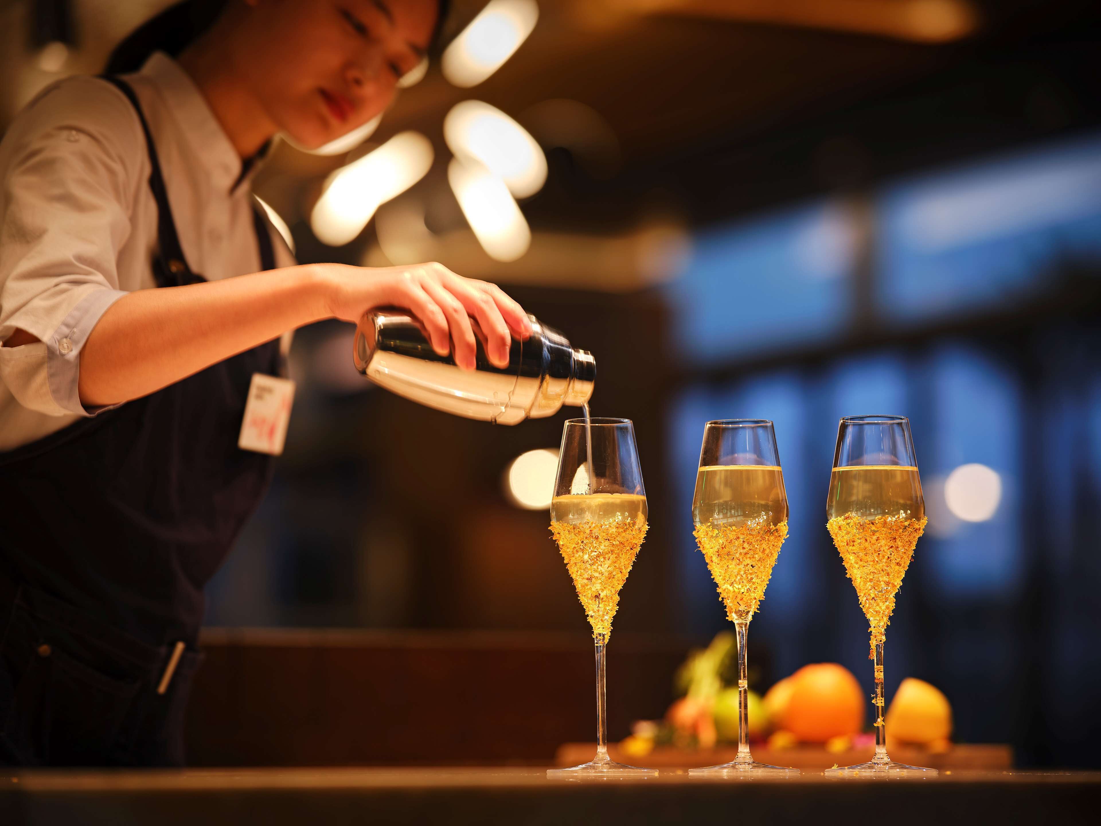 A bartender pouring a drink into two glasses with a golden garnish.