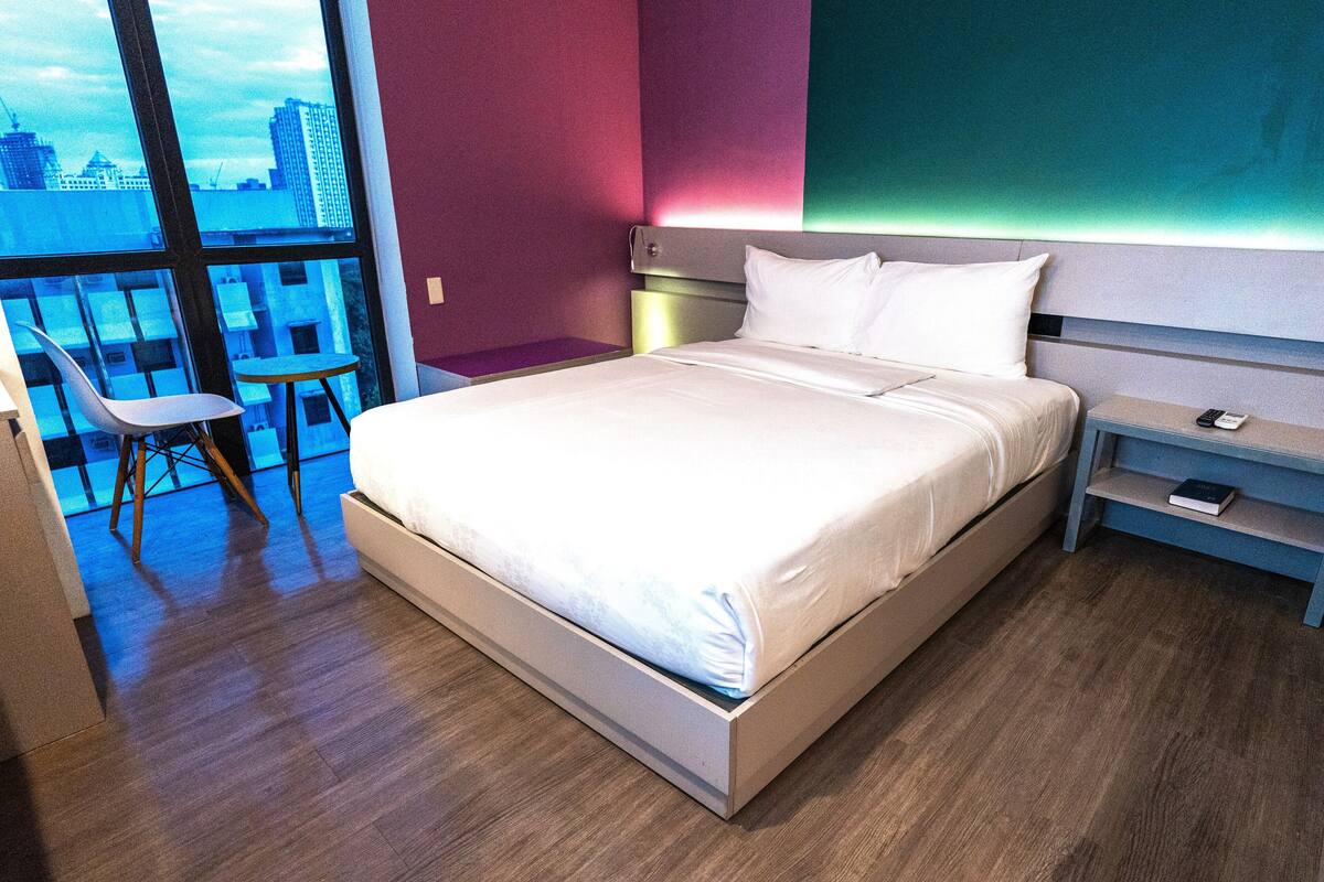 Quarto de hotel moderno com uma cama grande, uma mesa de cabeceira com telefone e uma cadeira.