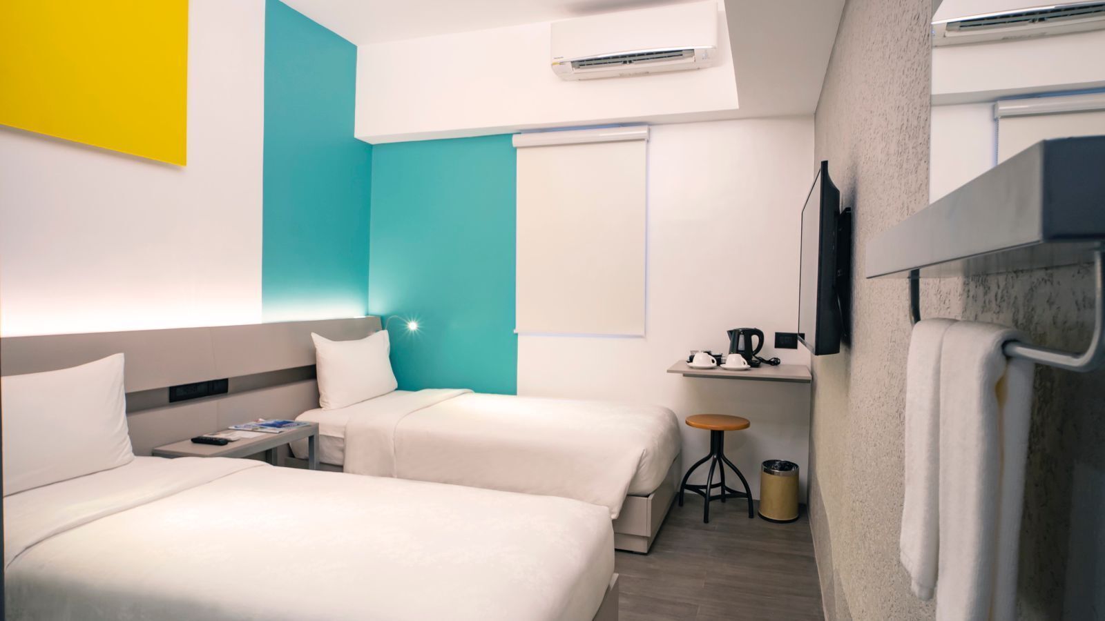 Habitación de hotel con dos camas, un escritorio pequeño con hervidor, un cubo de basura y un equipo de aire acondicionado.