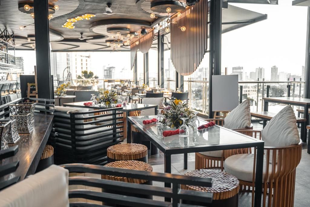 A modern restaurant with high ceilings, exposed beams, and large windows offering city views. Tables are set with white tablecloths, red napkins, and floral centerpieces.