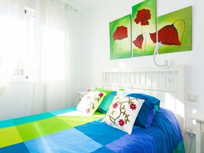 Een lichte slaapkamer met een bed, kleurrijk beddengoed en drie rode bloemschilderijen aan de muur.