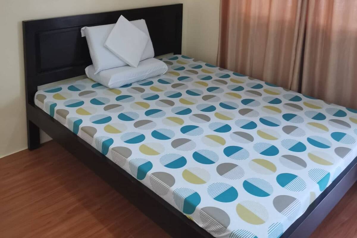 A single bed with a patterned bedspread, a dark headboard, and a window with curtains.