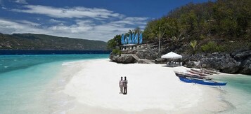 Una playa con orilla de arena blanca, algunas personas de pie y un bote azul cerca del agua.