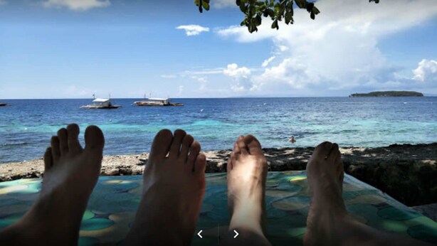 Escena de playa con los pies de una persona sobre una estera colorida, con vista al mar, barcos y una isla en la distancia.