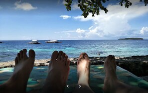 A beach scene with a person's feet on a colorful mat, overlooking the sea with boats and an island in the distance. - Island Front Bangcogon Resort and Restaurant (Oslob)