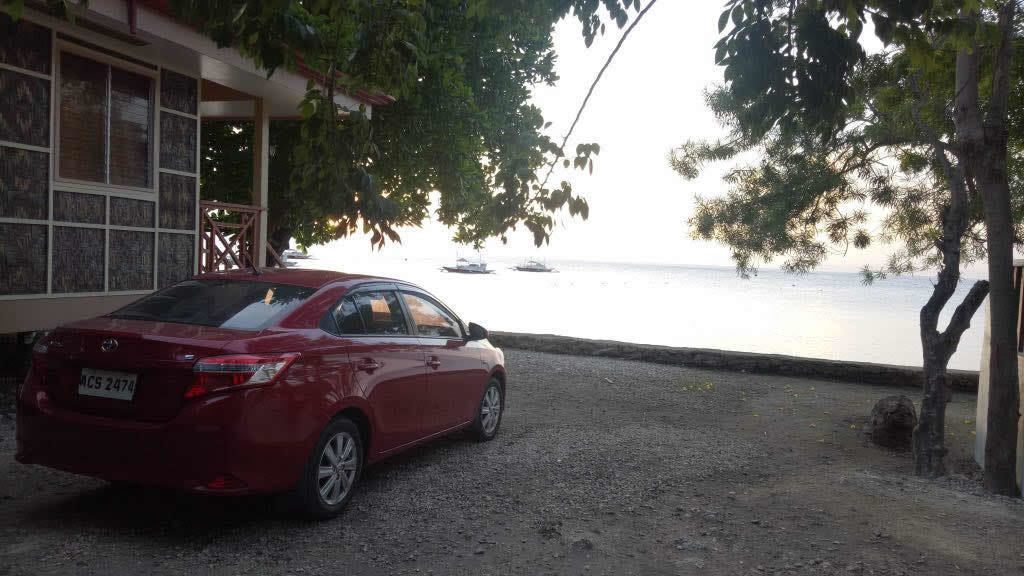 A red car parked on a gravel driveway with a building on the left and a view of the sea with boats in the background.