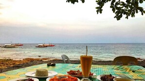 Una mesa servida con varios platos y una bebida, con vista a una playa donde se ven barcos a lo lejos.